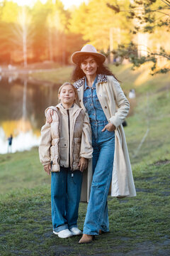 Smiling mother embraces young daughter beside tranquil autumn lake outdoors. Cheerful parent wears stylish hat and trench coat while supporting confident preteen girl on grassy riverbank.