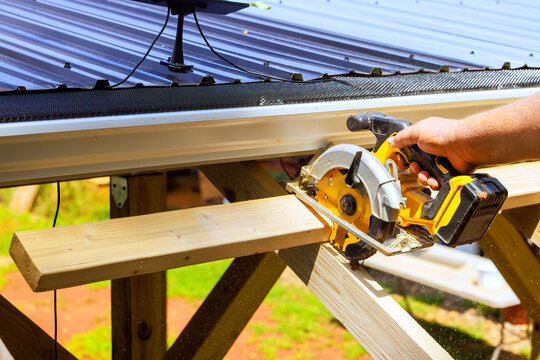 Worker cuts wooden beams plank with power saw while standing on construction site