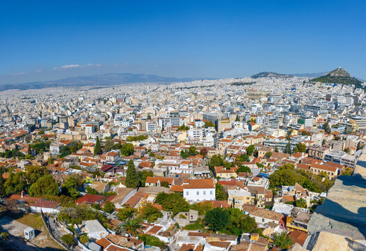 A wide elevated panoramic view of Athens, Greece, looking down from the Acropolis over the historic Plaka neighborhood towards Mount Lycabettus under a bright blue sky.