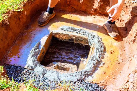 Worker works on concrete septic tank base inside dug hole in yard during construction water mud are present.