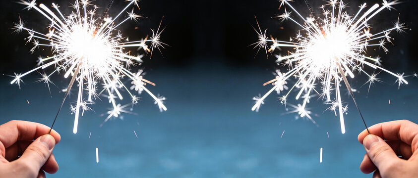Hands holding lit sparkling sparklers against a blurred blue night background with large copy space