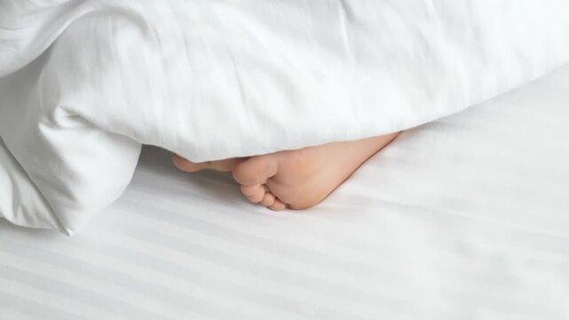 Feet slip under soft duvet in peaceful family bedroom, supporting cozy bedtime routine. Bare toes curl and settle beneath clean white sheets during quiet rest, then remain tucked under bedding.