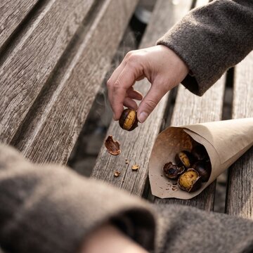 Close-up of a hand holding a steaming roasted chestnut over a wooden park bench next to a brown paper cone filled with freshly cooked autumn street food