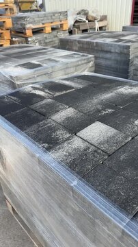 Pallets of dark grey concrete paving slabs wrapped in plastic film in storage. 