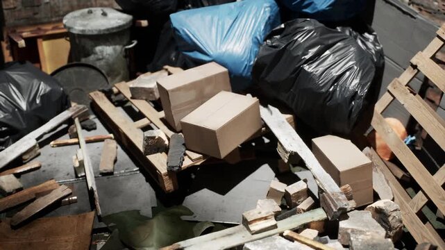 Messy collection of cardboard containers and waste bags resting on wooden pallets near old bricks and debris in a cluttered outdoor area during the bright daylight hours of a typical summer afternoon