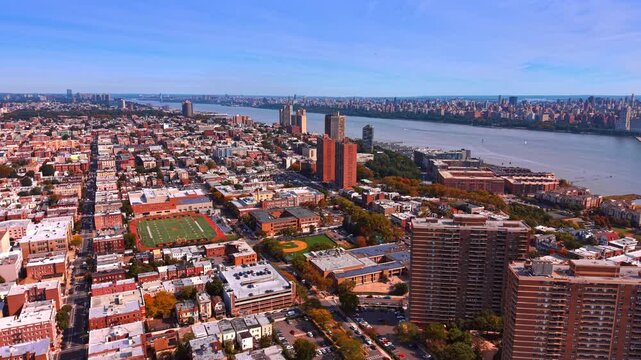 Residential blocks spread beside the Hudson in New Jersey. Rows of rooftops surround a fenced sports field below taller apartment buildings.