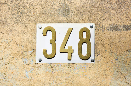 Number 348 brass house number on enamel plate screwed to cracked plastered wall, vintage metal digits with weathered texture, address sign detail for real estate, architecture