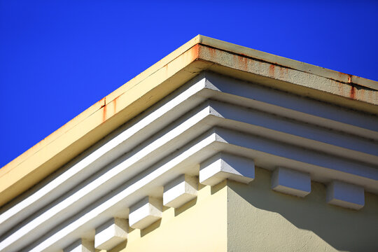 Classic building cornice corner with stepped moldings and dentils under clear skies, sunlit pastel facade with rusty stains, architecture detail for real estate, construction and design concepts