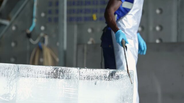 Low angle of large clear ice blocks in the foreground as an African worker in an apron and blue gloves drags a block with a steel ice hook across the factory floor