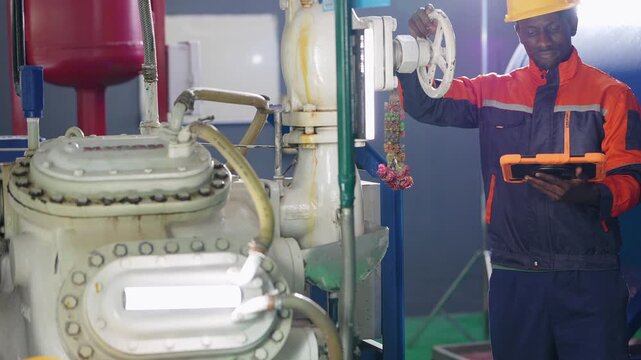 African engineer with an orange tablet raises his hand and adjusts the valve wheel over the compressor, a crouching colleague in a yellow hard hat below for maintenance