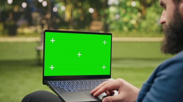 Businessman presents a laptop with green-screen chroma key against a blurred city park, providing a clean editable widescreen mockup for digital content and screen replacement outdoors.
