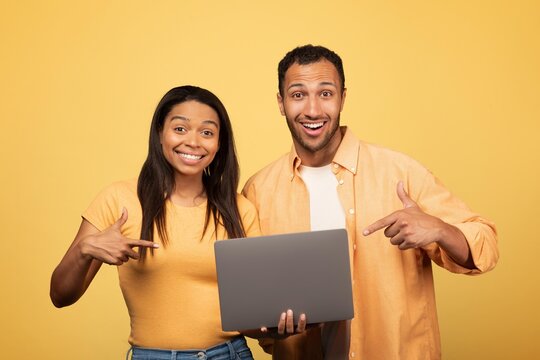 Smiling Black Man And Woman Pointing At Laptop Together, Expressing Joy And Excitement At A Workspace With Yellow Background. Engaged In Collaborative Activity, Focused On Business Concept.