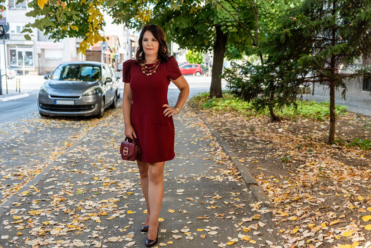 Elegant Woman in short purple dress and high heels, wearing beads necklace, carrying a small purple hand bag walking on a autumn street full of fallen leaves