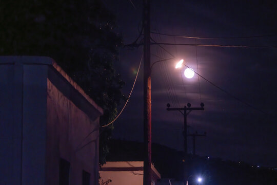 A bright full moon shines through wispy night clouds directly behind an electricity pole with power lines, illuminating the dark street and rooftops of a coastal village on Aegina Island, Greece.