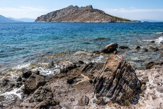 Clear turquoise waters lap against the rugged rocky shore near Perdika village on Aegina Island, offering a scenic view of Moni Island and distant sailboats in the Saronic Gulf.