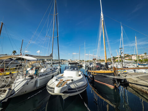 Sailboats, yachts and motorboats moored at Port du Lavandou on the French Riviera, showing crowded marina berths, tall masts, ropes and reflections on the still water under a blue sky.