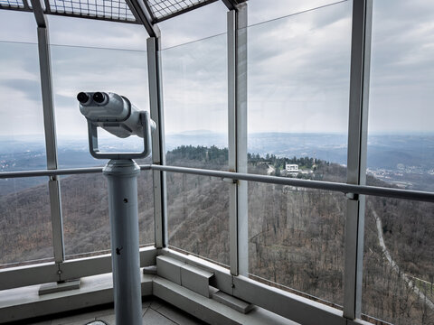 Fixed observation binoculars beside glass panels on the Avala Tower viewing platform, overlooking wooded slopes and a distant hazy panorama of the surrounding landscape in belgrade, serbia