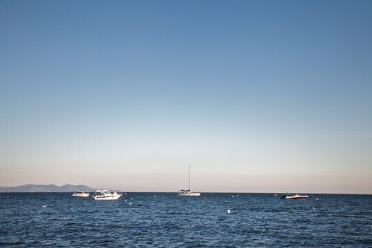 Leisure boats, including a sailboat and motorboats, lie at anchor on the Mediterranean Sea off the French Riviera, with mooring buoys and distant coastal hills visible.