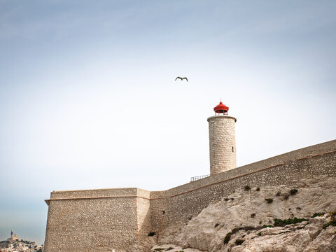 A stone lighthouse with a red lantern stands above fortified coastal walls and jagged rocks in Marseille, France, overlooking Mediterranean waters with rocky islets and distant land beneath a pale sky