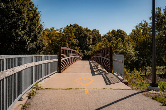 Paved pedestrian footbridge along a sunny park trail with steel handrails and surrounding green trees