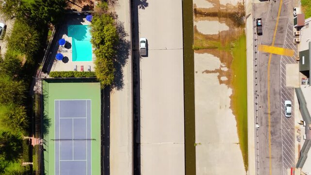 A white pickup truck drives along a dry concrete flood channel, top down drone view. Tennis court, turquoise pool with blue umbrellas, green algae stains and a parking lot on a sunny day.