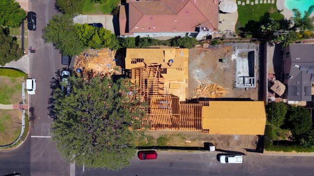 New house framing in Studio City, Los Angeles, seen top down. Construction worker on plywood deck, lumber piles and empty pool shell. Red tile roofs, backyard pools and parked cars on a sunny street.