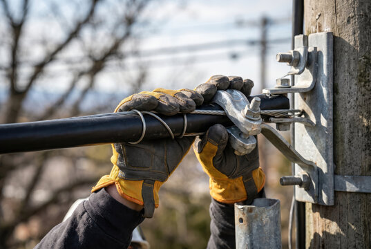 Gloved hand securing tension clamp to thick black cable on utility pole hardware, with blurred outdoor background.