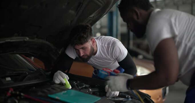Two mechanics inspecting car air conditioning system with manifold gauge and flashlight