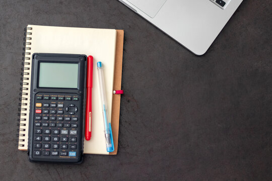 Top view of open notebook with calculator, pens, and laptop on dark desk