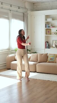 Young woman dances joyfully at home, raising her arms and hopping across a sunlit living room. Her energetic movement celebrates happiness, self-expression, wellness and active home leisure.