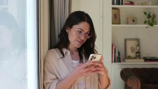 Young woman uses a smartphone at home, typing and scrolling beside a window with sheer curtains. She looks outside with a thoughtful smile, then returns to texting in her softly lit modern apartment.