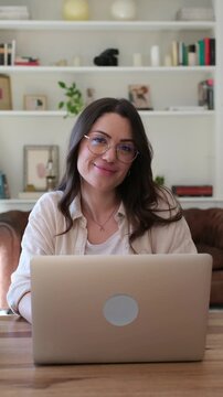 Woman works on a laptop at home, smiling at the camera and resting her chin on her hand. Young freelancer in glasses sits at a table in a home office, conveying remote work and online communication.