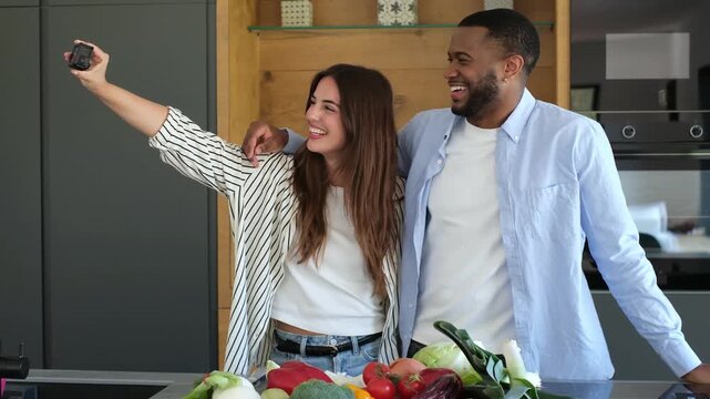 Couple records a cooking vlog in the kitchen as the woman holds a camera and her partner hugs her behind fresh vegetables. Their filming captures laughter and healthy food content for social media.