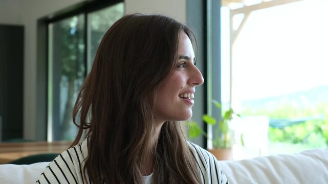 Young woman smiles and looks out the window while sitting on a sofa at home. Her relaxed side profile in a bright living room conveys wellness, quiet leisure, mindfulness, and domestic lifestyle.