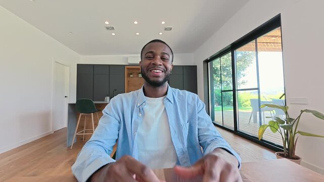 Young Black man talks and gestures during a video call from home, looking into the camera with wireless earbuds. He smiles at a wooden table in a bright kitchen living space with sunny trees outside.