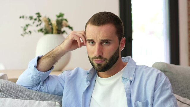 Young man sits thinking on a couch at home, resting his head in his hand and rubbing his temple. He shifts his gaze toward the camera in a bright living room, appearing pensive and contemplative.