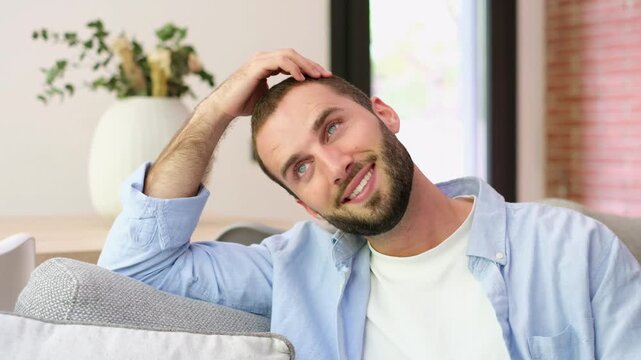 Young man smiles and relaxes on a sofa at home, resting his head on one hand and looking upward thoughtfully. Casual living room lifestyle clip conveys leisure, wellbeing, daydreaming and comfort.