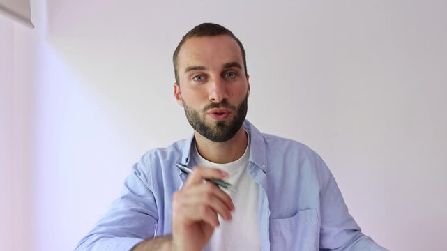 Young man waves hello, speaks and gestures to the camera during a video call at home. He holds a pen, explains an idea and taps his temple, supporting online communication, vlog or learning content.