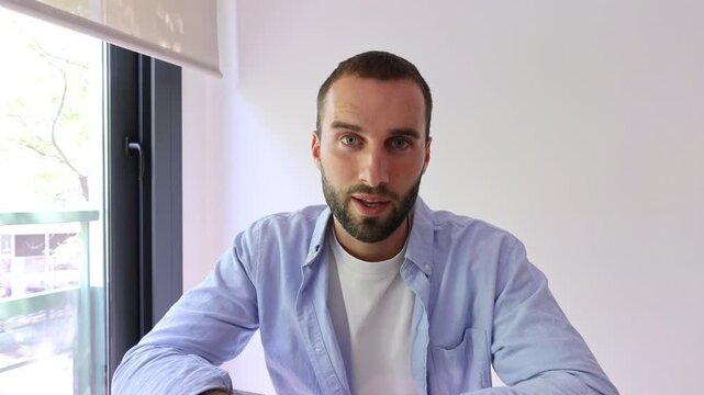 Young man talks to the camera with a warm smile and hand gestures during an online video call at home. He communicates directly with viewers in a bright room for vlog, interview, or learning content.