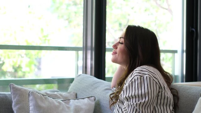 Woman relaxes on a couch, smiling and thinking as she looks out the window in a bright living room. Her expression becomes contemplative while she enjoys a quiet moment and watches trees in daylight.