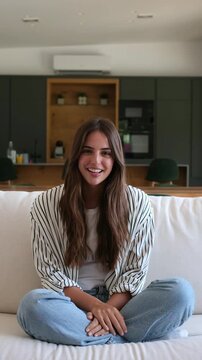 Woman talks to the camera during a video call at home, gesturing while seated cross-legged on a couch. She appears in a bright living room with a blurred kitchen behind her, explaining her message.