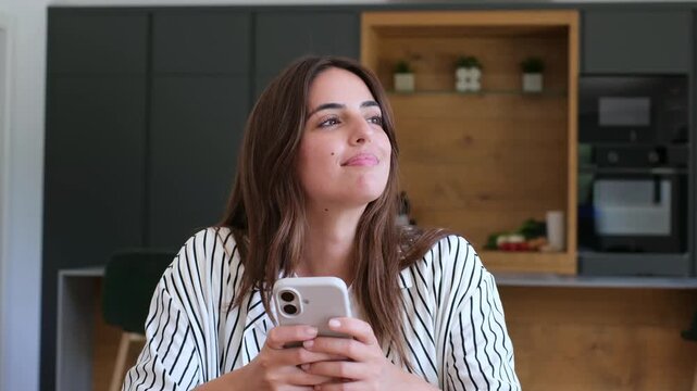Young woman uses a smartphone and smiles while typing messages in her home kitchen. She looks up thoughtfully, then returns to mobile communication on her phone with a cheerful expression by dark.