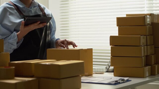 A seller uses a tablet beside stacked parcels in a packing area. Cardboard boxes and digital tools illustrate ecommerce order management, small business shipping, and preparation for parcel delivery.