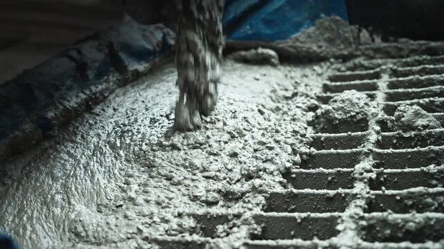 Close-up pouring of fresh concrete mix over metal grate in construction setting, wet textured cement flowing from chute and filling grating surface under soft industrial light