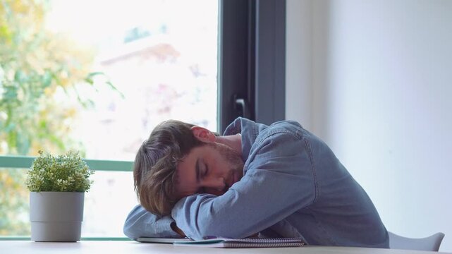 Young man types on a laptop, then closes it and rests his head on the desk by a window, showing work stress, fatigue and burnout. Static medium shot captures exhaustion during a demanding workday.