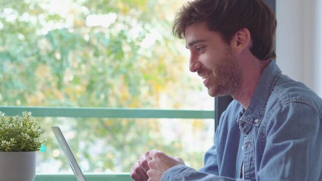Young man works from home at a desk, talking and gesturing during a video call on his laptop. He holds a pen and communicates in a bright home office beside a window with green trees outside.