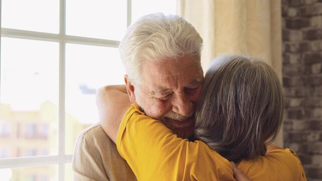 Senior couple hug tightly and smile together at home beside a bright window. Elderly man and woman share love, affection, romance and companionship, expressing a lasting marriage relationship.