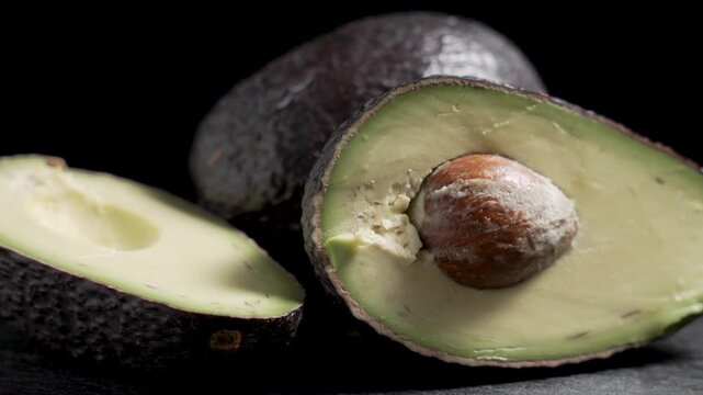 Low angle close-up of two avocado halves, one with the pit, and a whole avocado on dark slate against a black background as the camera slowly approaches.