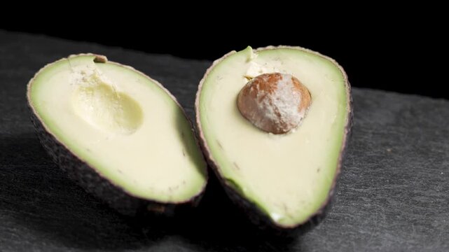 Two fresh avocado halves, one with the brown pit, rest side by side on a dark slate board while the camera slowly circles around them.