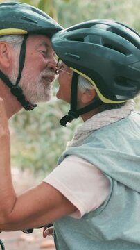Senior couple wearing cycling helmets smile, kiss and share affection outdoors in soft daylight. Active older man and woman enjoy fitness, romance and a healthy retirement lifestyle amid foliage.